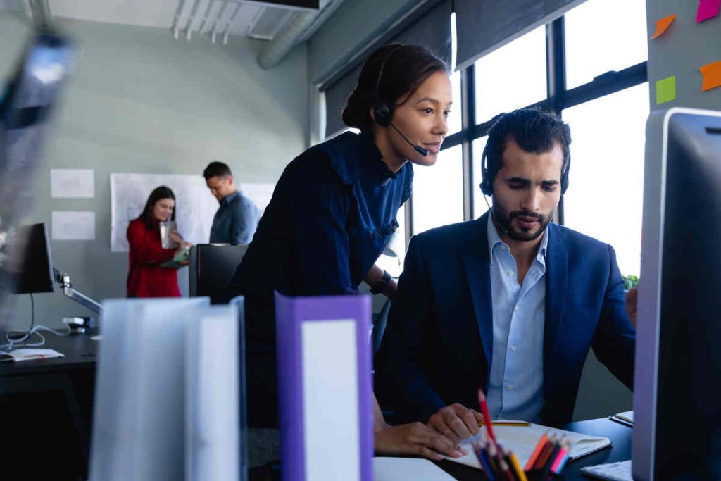 Caucasian man and mixed race woman wearing headset, using a desktop computer, with other colleagues working in the background.