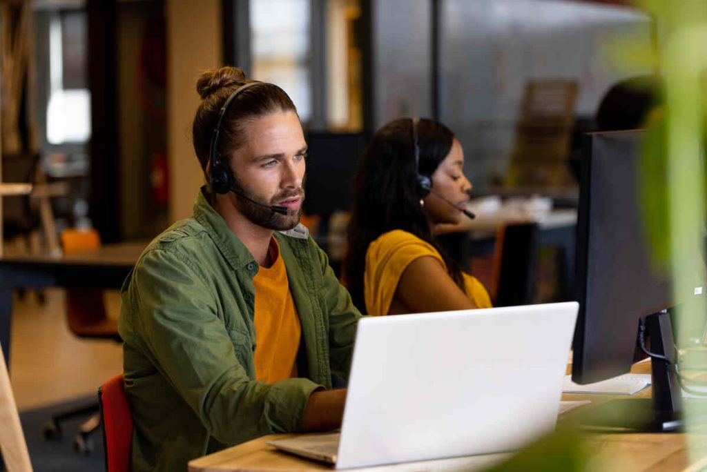 Diverse male and female creative colleague in phone headsets using computers in casual office. Casual office, communication, business and work, unaltered.