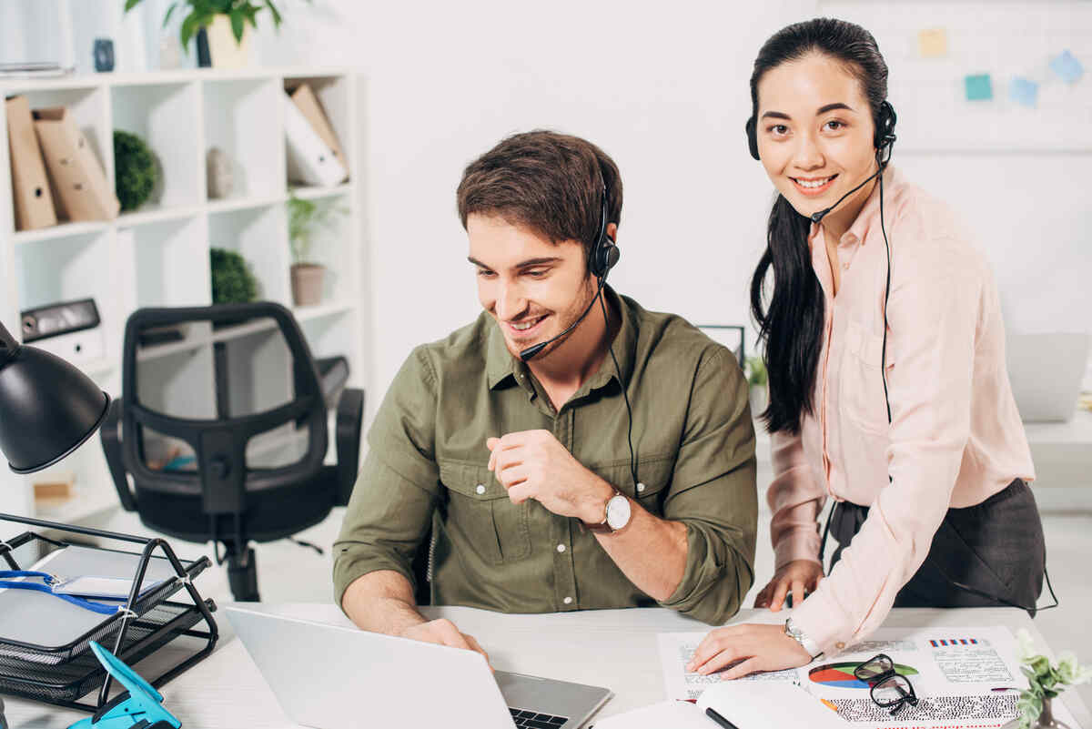 smiling call center operator working at laptop and pretty coworker looking at camera in office